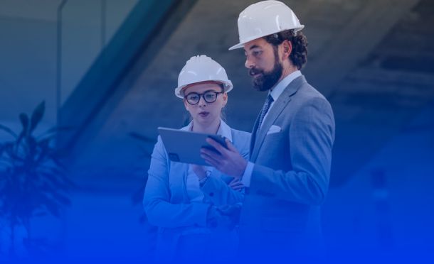homem e mulher, com capacete de EPI e tablete na mão, em ambiente de empresa de engenharia